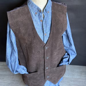 May include: A dark brown suede vest featuring a V-neckline, four front buttons, and two lower pockets, layered over a light blue long-sleeved button-down shirt with a textured fabric finish and brown buttons on a mannequin torso.