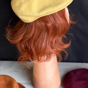 Puede incluir: Un boina amarilla se muestra sobre una cabeza de maniqu&iacute; con cabello casta&ntilde;o rojizo. Tambi&eacute;n se ven un sombrero fedora marr&oacute;n, una boina burdeos y dos hojas de oto&ntilde;o. El fondo es negro.