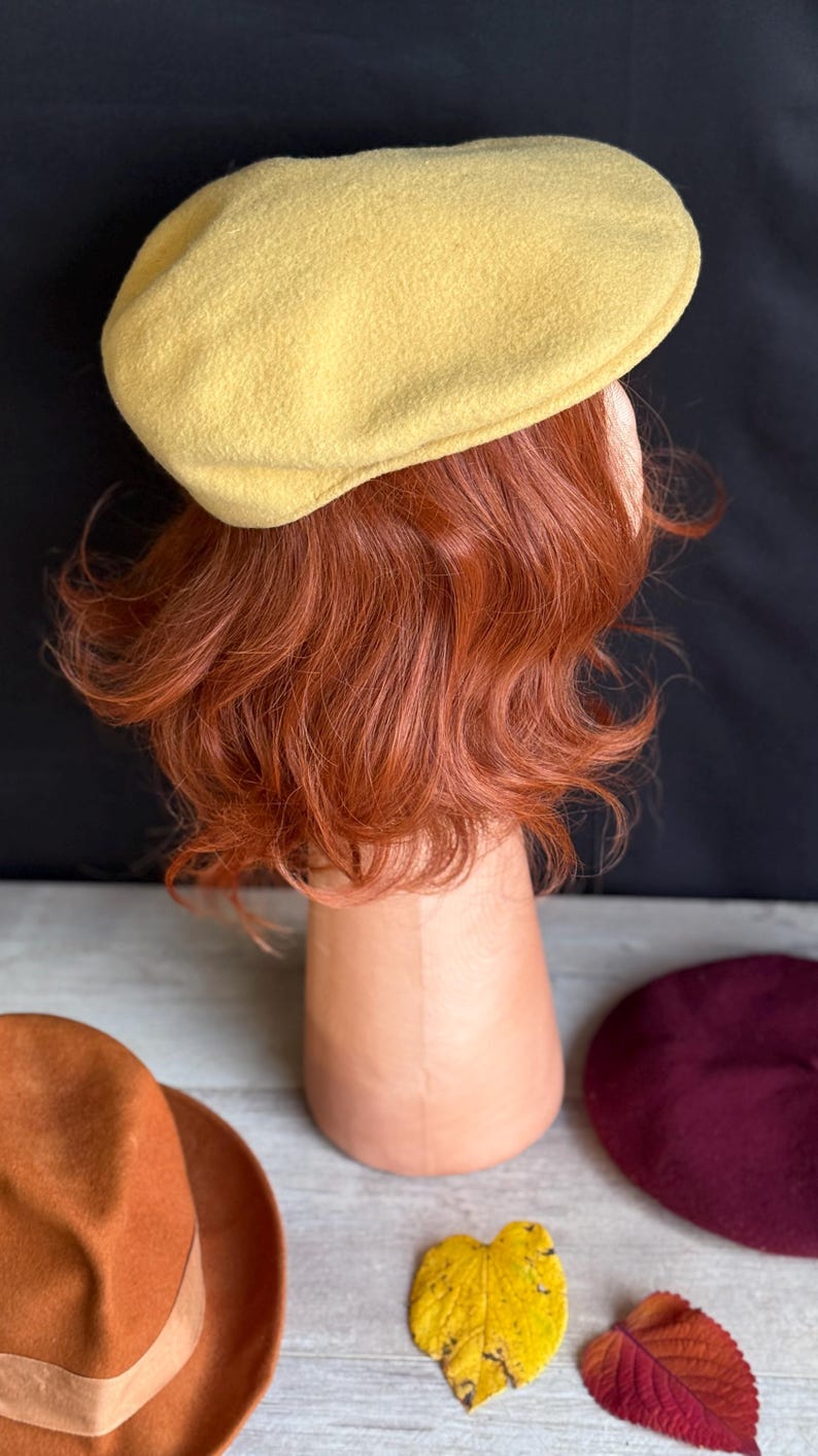 Puede incluir: Un boina amarilla se muestra sobre una cabeza de maniqu&iacute; con cabello casta&ntilde;o rojizo. Tambi&eacute;n se ven un sombrero fedora marr&oacute;n, una boina burdeos y dos hojas de oto&ntilde;o. El fondo es negro.