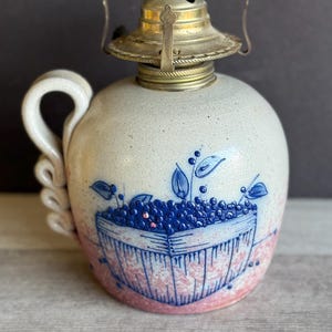 May include: A ceramic oil lamp with a round body and a handle. The lamp features a hand-painted design of a basket filled with blueberries, with blue leaves and pink accents. The top is brass-colored.