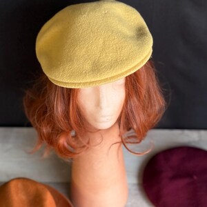 Puede incluir: Un boina de color amarillo mostaza se encuentra sobre una cabeza de maniqu&iacute; con cabello casta&ntilde;o rojizo. Un sombrero fedora marr&oacute;n y una boina burdeos son visibles debajo. Una hoja amarilla y una hoja roja tambi&eacute;n est&aacute;n en la imagen. El fondo es negro.
