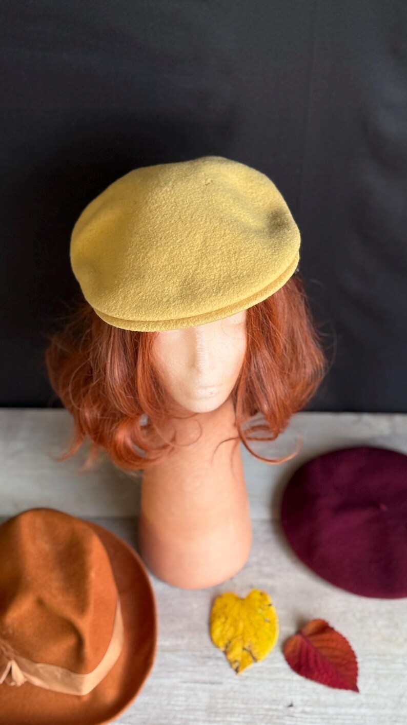 Puede incluir: Un boina de color amarillo mostaza se encuentra sobre una cabeza de maniqu&iacute; con cabello casta&ntilde;o rojizo. Un sombrero fedora marr&oacute;n y una boina burdeos son visibles debajo. Una hoja amarilla y una hoja roja tambi&eacute;n est&aacute;n en la imagen. El fondo es negro.
