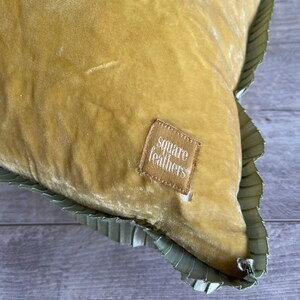 K&ouml;nnte beinhalten: Ein senfgelbes Samtkissen mit gr&uuml;nem R&uuml;schenbesatz. Das Kissen hat ein kleines braunes Etikett mit der Aufschrift "square feathers".