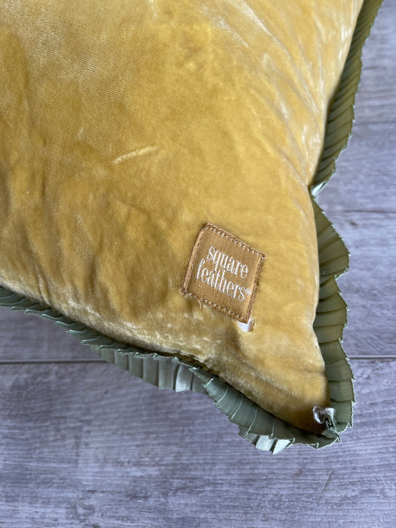 K&ouml;nnte beinhalten: Ein senfgelbes Samtkissen mit gr&uuml;nem R&uuml;schenbesatz. Das Kissen hat ein kleines braunes Etikett mit der Aufschrift "square feathers".