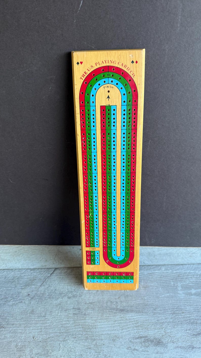 May include: Wooden cribbage board with red, green, and blue tracks. The board has the text "THE U.S. PLAYING CARD CO." and a start and finish point. The board is rectangular and has numerous holes for pegs.