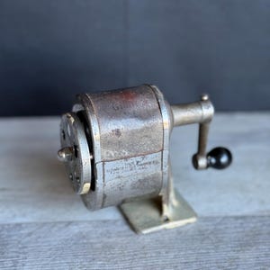 May include: A vintage metal pencil sharpener with a hand crank and a black knob. The cylindrical body features the text Giant No. 2 and Made by Automatic Pencil Sharpener Co. Chicago, Ill. The device has a weathered finish and a circular front plate.