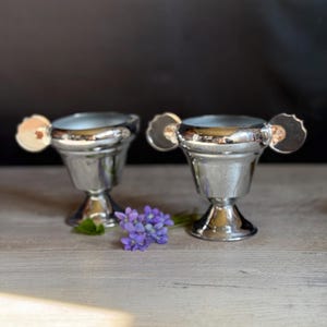May include: Two shiny silver egg cups with decorative handles. The egg cups are on a light-colored wooden surface, with a small bunch of purple flowers and green leaves between them. The background is a dark gray.