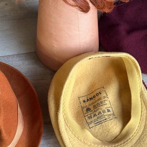 Puede incluir: Una colecci&oacute;n de sombreros y un soporte para pelucas. Los sombreros incluyen una gorra Kangol amarilla, un fedora color &oacute;xido y una boina burdeos. Una peluca roja se encuentra sobre un soporte de madera para pelucas.