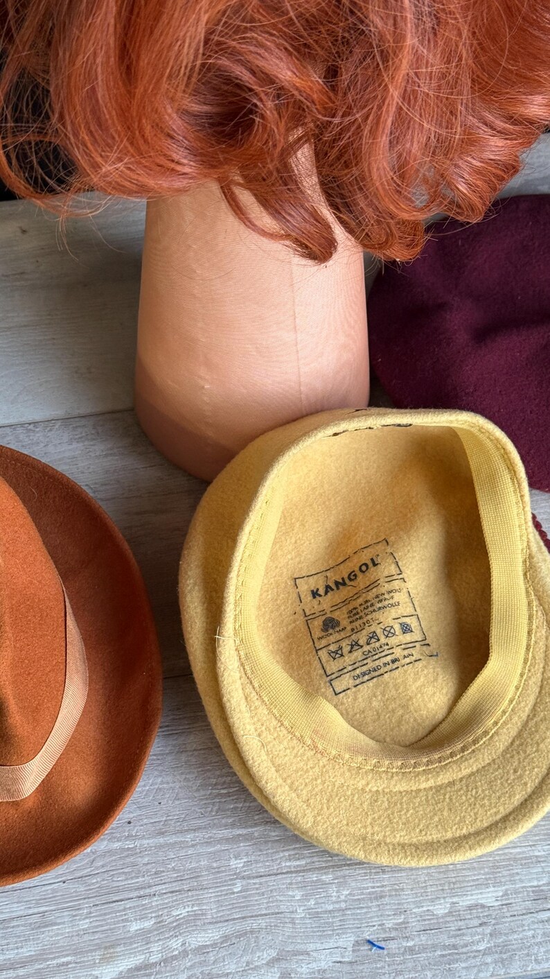 Puede incluir: Una colecci&oacute;n de sombreros y un soporte para pelucas. Los sombreros incluyen una gorra Kangol amarilla, un fedora color &oacute;xido y una boina burdeos. Una peluca roja se encuentra sobre un soporte de madera para pelucas.