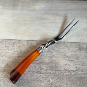May include: A vintage carving fork with a two-tine stainless steel head and a handle made of amber and dark brown material. The handle is accented with a silver-colored metal ferrule.