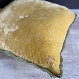 K&ouml;nnte beinhalten: Ein senfgelbes Samtkissen mit gr&uuml;nem R&uuml;schenbesatz. Das Kissen hat ein kleines braunes Etikett mit dem Text "square feathers".