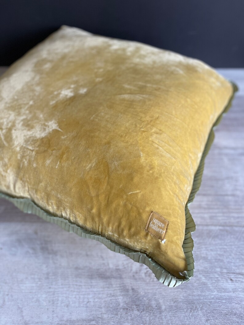 K&ouml;nnte beinhalten: Ein senfgelbes Samtkissen mit gr&uuml;nem R&uuml;schenbesatz. Das Kissen hat ein kleines braunes Etikett mit dem Text "square feathers".