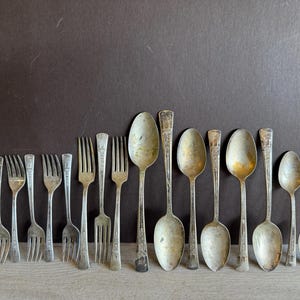 May include: A collection of vintage silverware, including forks and spoons, with an aged silver finish. The silverware is arranged in a row on a light brown surface.