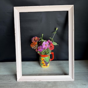 May include: A white rectangular picture frame with distressed edges surrounds a colorful floral arrangement. The flowers are in a small, decorative pitcher with a yellow base and fruit designs. The background is black.
