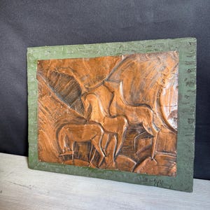 May include: A rectangular copper wall plaque featuring a relief design of two horses standing in a landscape. The central copper panel is framed by a textured, dark green border. The metal surface shows visible hammer marks and metallic tones.