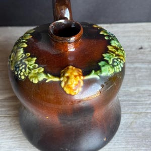 May include: A brown ceramic pitcher with a handle and a small spout. The pitcher is decorated with green and yellow grape vines and a yellow face. The pitcher has a bulbous body and sits on a wooden surface.