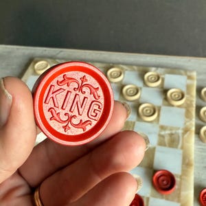 May include: A red checker piece with the word "KING" in white lettering, held above a checkerboard. The checkerboard has a white and tan checkered pattern. Other checker pieces are visible.