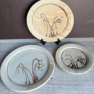 May include: Three handmade ceramic plates in varying sizes. The plates are beige with brown abstract designs. The largest plate is round, the middle plate is oval, and the smallest is round. The plates are displayed on a wooden surface.