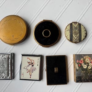 May include: A collection of seven vintage compact mirrors and cases. The top row features three round compacts in gold, black, and patterned finishes. The bottom row includes a silver mesh case, a dancer illustration, a black case, and a floral print.