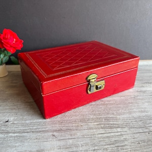 May include: A red rectangular box with a gold clasp and a geometric pattern on the lid. The box is made of a leather-like material and has gold trim. A red rose is in the background.