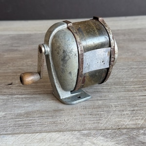 May include: Vintage metal pencil sharpener with a silver-colored body, a wooden handle, and a cylindrical blade housing. The sharpener shows signs of age and use, with visible wear and rust.