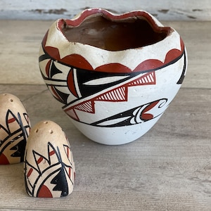 May include: Two small white ceramic salt and pepper shakers with red, black, and white geometric designs. A larger white ceramic bowl with a similar design is in the background.