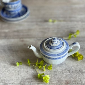 May include: A blue and white porcelain teapot with a floral design. The teapot is sitting on a light wood surface with green leaves scattered around it.
