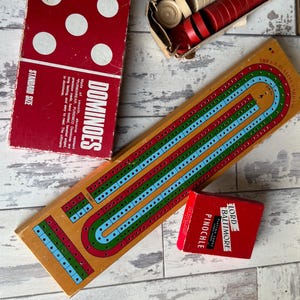 May include: A collection of vintage board games, including a red dominoes box with white dots, a wooden cribbage board with blue, green, and red lines, and a red box of "Pinochle" playing cards. Also, a box of red and beige game pieces.