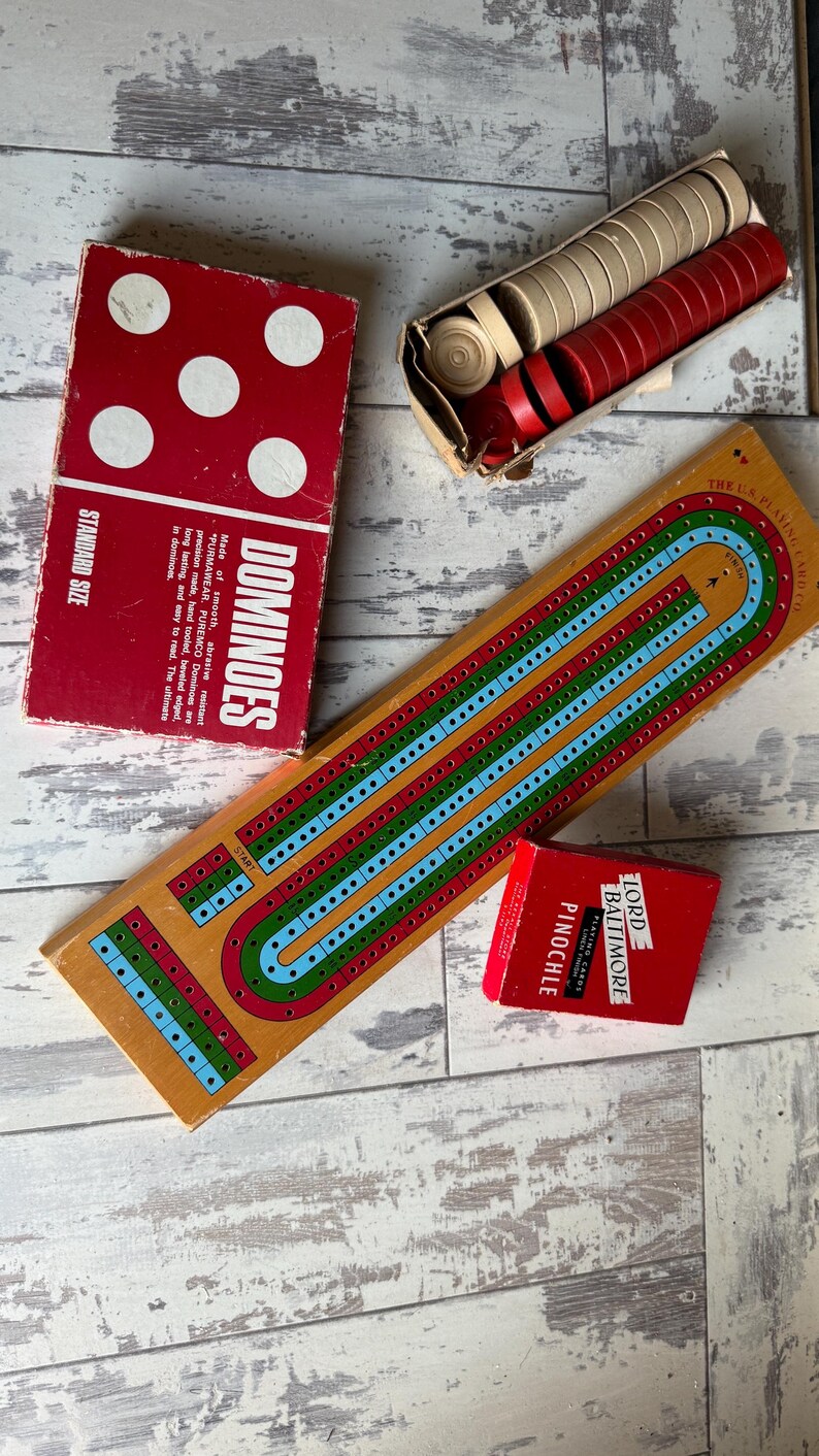 May include: A collection of vintage board games, including a red dominoes box with white dots, a wooden cribbage board with blue, green, and red lines, and a red box of "Pinochle" playing cards. Also, a box of red and beige game pieces.
