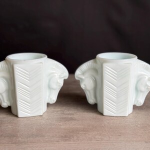 May include: Two white art deco candle holders. Each holder features a central column with a chevron design and is flanked by two horse head sculptures. The candle holders are made of a glossy white material and sit on a wooden surface.