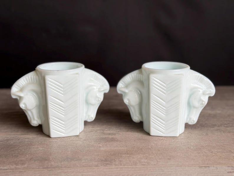 May include: Two white art deco candle holders. Each holder features a central column with a chevron design and is flanked by two horse head sculptures. The candle holders are made of a glossy white material and sit on a wooden surface.