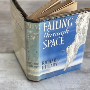May include: An open vintage book titled "Falling through Space" by Richard Hillary. The book cover is light blue with white text and a stylized illustration. The pages are aged, and the book is slightly worn.