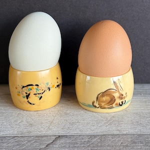 May include: Two yellow egg cups with painted designs. One cup has a floral design and the other has a rabbit design. A white egg is in the floral cup and a brown egg is in the rabbit cup.