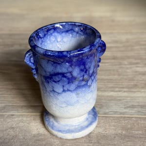 May include: A small ceramic vessel featuring a crystalline glaze that transitions from a deep cobalt blue at the rim to a pale, icy white at the base. The piece has a flared opening, a pedestal foot, and two small, decorative side handles.