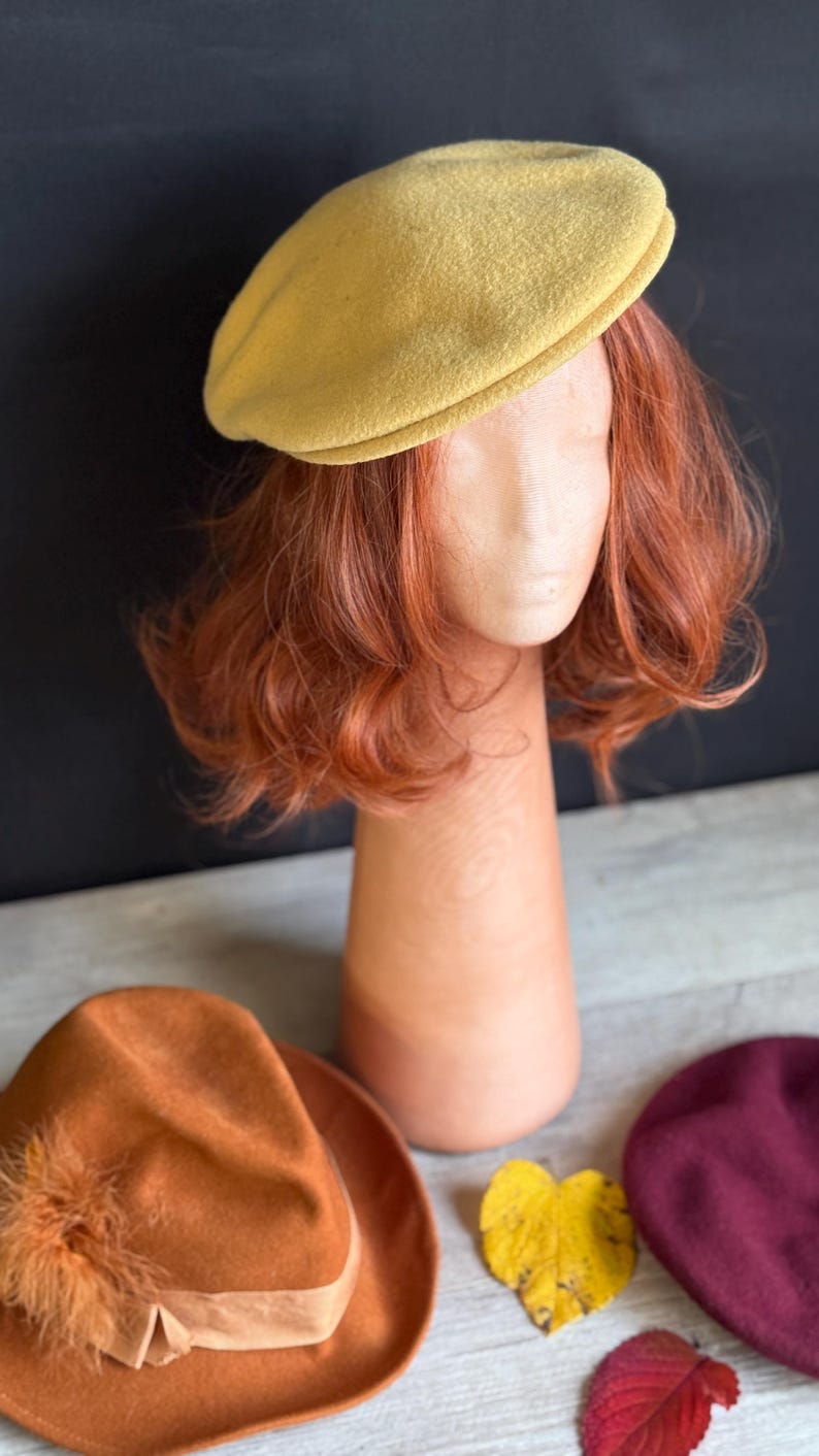 Puede incluir: Un boina amarillo mostaza se muestra sobre una cabeza de maniqu&iacute; con cabello casta&ntilde;o rojizo. Tambi&eacute;n se exhiben un sombrero fedora naranja con un adorno de plumas y una boina burdeos. Tambi&eacute;n hay una hoja amarilla y una hoja roja.