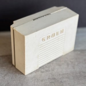 May include: A vintage off-white Gruen watch box. The rectangular box has a hinged lid and a grooved front panel with the brand name embossed. The box is set against a gray wooden surface and a dark background.