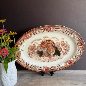 May include: Oval serving platter with a turkey illustration in shades of brown, green, and pink. The rim features a brown border with floral and fruit accents. The platter is on a black stand, with a white pitcher of flowers to the left.