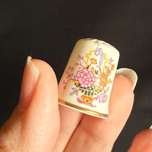 May include: A vintage thimble with a floral design. The thimble is off-white with a gold rim and features a colorful arrangement of pink, yellow, and blue flowers. The thimble is held between fingers against a dark background.