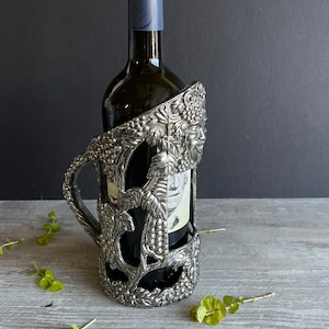 Puede incluir: Porta botellas de vino de plata con diseño de vid. El porta botellas tiene forma de vid con uvas y hojas. Una botella de vino está en el porta botellas.