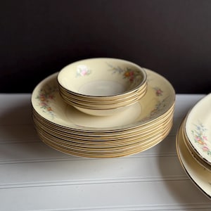 May include: A stack of cream-colored vintage china plates and bowls. Each piece features a delicate floral pattern in pink, yellow, and blue, with gold trim. The plates and bowls are stacked on a white surface against a black background.