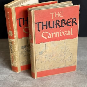 May include: Two vintage copies of "The Thurber Carnival" by James Thurber. The books have red covers with black and white text and illustrations. The spines are also red with the author's name. The books are on a light wood surface.