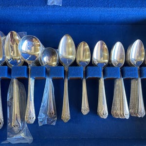 May include: A collection of silver-toned metal spoons arranged in a blue velvet-lined storage case. The set includes various sizes of spoons, some wrapped in clear plastic, all featuring ornate decorative detailing on the handles and bowls.