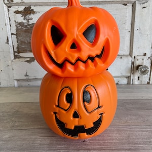 May include: Two orange plastic jack-o'-lantern decorations stacked on top of each other. The top jack-o'-lantern has a mean expression with black eyes and a wide, toothy grin. The bottom jack-o'-lantern has a friendly expression with large, round eyes and a wide, toothy grin.