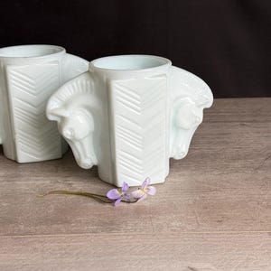 May include: Two white art deco vases, each featuring a horse head design on the sides. The vases have a geometric pattern on the front and are displayed on a wooden surface. A small purple flower lies in front of the vases.