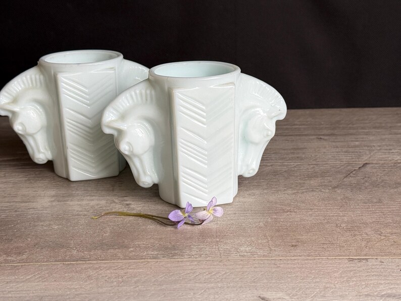 May include: Two white art deco vases, each featuring a horse head design on the sides. The vases have a geometric pattern on the front and are displayed on a wooden surface. A small purple flower lies in front of the vases.