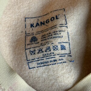 Puede incluir: Primer plano del interior de un sombrero Kangol beige con una etiqueta azul. La etiqueta incluye el logotipo de Kangol, la marca de lana y texto que indica que el sombrero est&aacute; hecho de 100% lana virgen pura y dise&ntilde;ado en Gran Breta&ntilde;a. El interior del sombrero es un material suave similar al fieltro.
