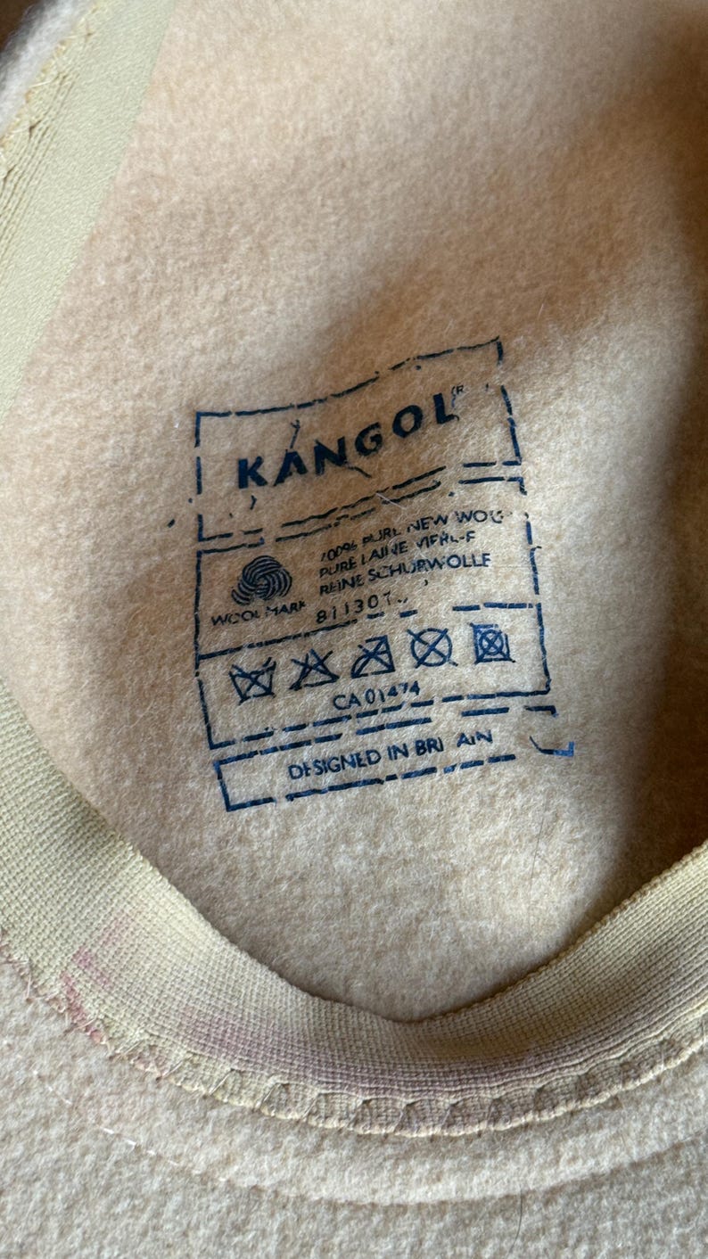 Puede incluir: Primer plano del interior de un sombrero Kangol beige con una etiqueta azul. La etiqueta incluye el logotipo de Kangol, la marca de lana y texto que indica que el sombrero est&aacute; hecho de 100% lana virgen pura y dise&ntilde;ado en Gran Breta&ntilde;a. El interior del sombrero es un material suave similar al fieltro.