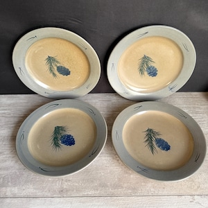 May include: Four oval-shaped ceramic plates with a light tan center and a blue-gray rim. Each plate features a painted pine cone and branch design in blue and green. The plates are arranged on a light-colored surface.