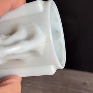 May include: A white, cylindrical container with a detailed relief design. The container has a smooth, glossy finish and a slightly curved rim. The relief appears to depict a classical figure or scene. The container is held by a person.