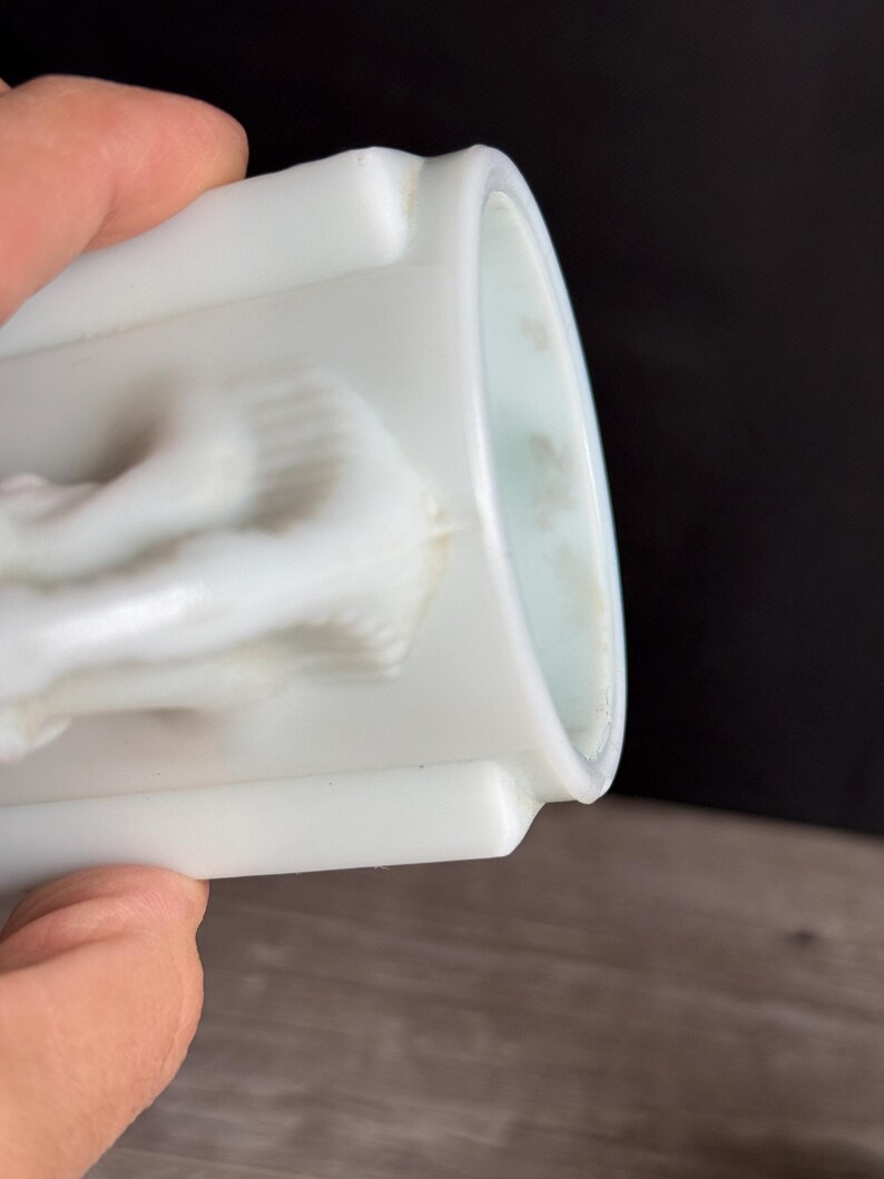 May include: A white, cylindrical container with a detailed relief design. The container has a smooth, glossy finish and a slightly curved rim. The relief appears to depict a classical figure or scene. The container is held by a person.